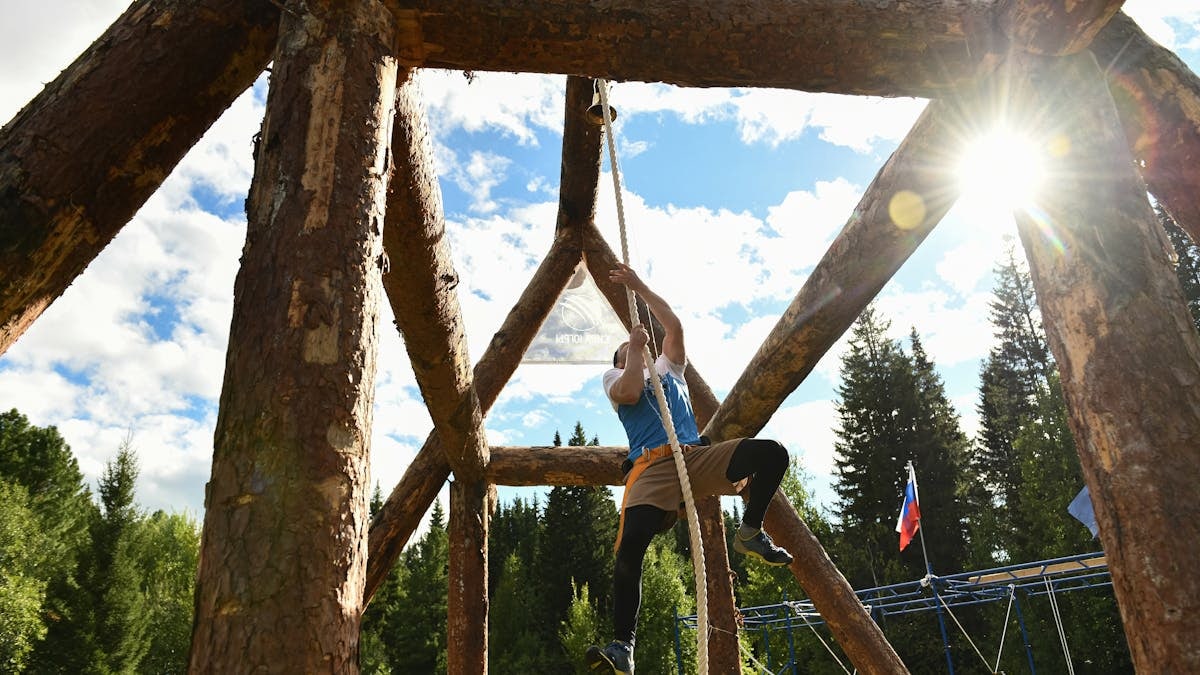 Adult man climbs rope on a wooden obstacle structure during a sunny day.